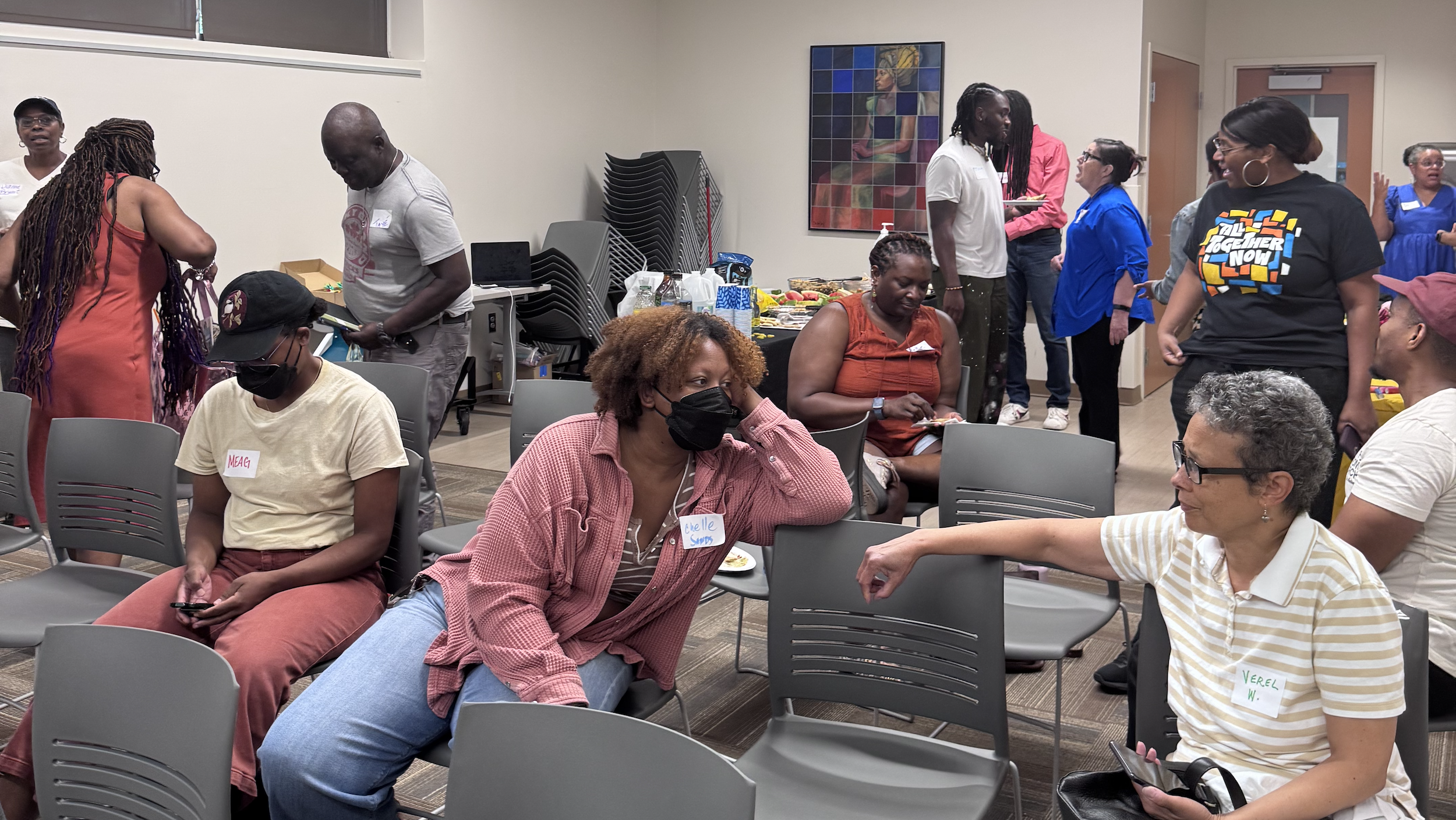 People attend an in person onboarding session at the West End library. Some folks sitting in chairs chatting, or reviewing the materials. Others are standing in the background talking.